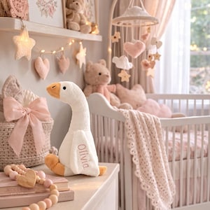 May include: A nursery scene with a white crib, a plush goose toy, and decorative elements in soft pink and beige tones. The goose toy has the name "Olivia" embroidered on it. A mobile with stars and hearts hangs above the crib.