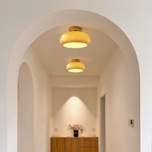 Puede incluir: Dos luces de techo amarillas y de madera iluminan un pasillo. Las luces tienen una forma redondeada, como de cuenco, con una base de madera. El pasillo cuenta con un mueble de madera y un jarrón con flores secas.
