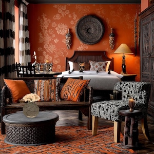Könnte beinhalten: Ein Schlafzimmer-Interieur mit afrikanischem Thema. Der Raum verfügt über einen dunklen Holzbettenrahmen, eine Bank mit gemusterten Kissen und einen gemusterten Teppich. Die Wände sind orangefarben mit floralen Mustern, und der Raum ist mit Holzmasken und einem runden Wandbehang dekoriert.
