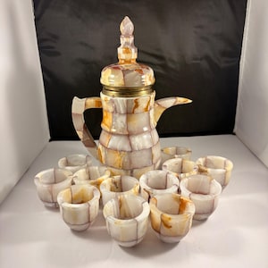 May include: A decorative onyx coffee set featuring a tall, lidded coffee pot and several small cups. The set is made of white, orange, and brown onyx tiles. The coffee pot has a brass-colored band around the middle.