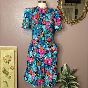 May include: A vintage floral dress with a vibrant pattern of pink, blue, and orange flowers on a black background. The dress features short sleeves, a cinched waist, and a large bow detail. The dress is displayed on a mannequin.