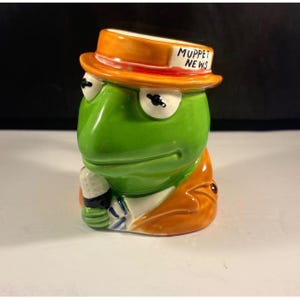 Puede incluir: Una taza de cerámica con forma de cabeza de rana verde que lleva un sombrero naranja con el texto MUPPET NEWS impreso en el ala. El personaje viste una chaqueta naranja sobre una camisa blanca y sostiene un micrófono blanco y negro en la mano.