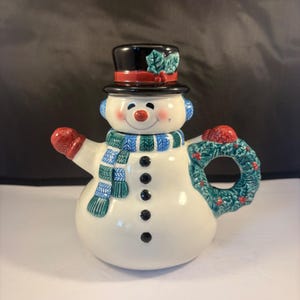 Könnte beinhalten: Eine Schneemann-Teekanne aus Keramik mit einem schwarzen Zylinder samt Stechpalme, einem blau-grün gestreiften Schal und einem Henkel in Form eines festlichen Kranzes mit roten Beeren. Der Schneemann hat eine Karottennase und fünf schwarze Knöpfe.