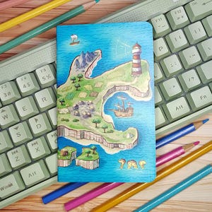 May include: A colorful notebook featuring a whimsical island map illustration with a lighthouse, sailing ships, and small villages. The notebook is open on a light green keyboard, with colored pencils in the background.