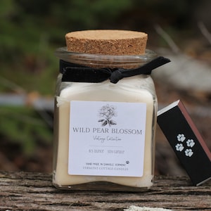 May include: A glass jar candle with a cork lid and a black ribbon tied around it. The label reads "WILD PEAR BLOSSOM Vintage Collection 8.5 ounce soy candle HAND MADE IN SHINNECOCK VERMONT VERMONT COTTAGE CANDLES".