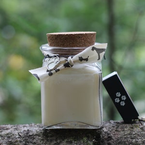 May include: A glass jar with a cork lid and a white ribbon tied around it. The ribbon has a silver paw print charm and a black and white dog print. The jar is filled with a white substance and is sitting on a tree branch.