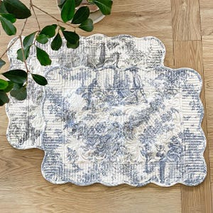 Op de afbeelding: Twee gewatteerde placemats met een geschulpte rand. De placemats hebben een blauw-wit toile de Jouy-patroon dat een pastorale scène uitbeeldt. De placemats liggen op een houten oppervlak, met een groene plant in de linkerbovenhoek.