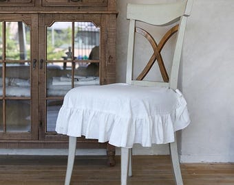 May include: A white chair with a ruffled seat cover. The chair has a light-colored wooden frame with an X-shaped back design. A wooden cabinet is in the background.
