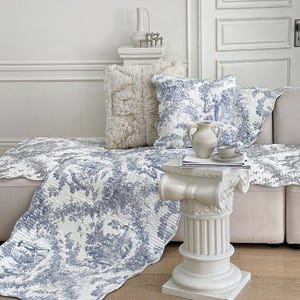 Op de afbeelding: Een crèmekleurige bank is versierd met een blauw-wit toile de Jouy-patroon quilt en bijpassende kussens. Een donzig, crèmekleurig kussen en een kleine witte decoratieve zuil met een vaas en kopje completeren de scène.