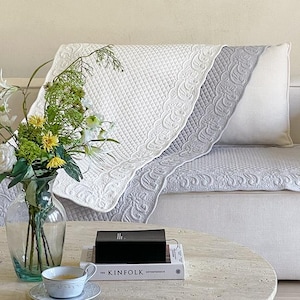 Könnte beinhalten: Eine weiße und graue gesteppte Sofahusse mit dekorativem Blumenrand ist über ein weißes Sofa drapiert. Eine Vase mit Blumen steht auf einem Marmortisch, zusammen mit einer Teetasse und einem Buch mit dem Titel "The Kinfolk Entrepreneur".