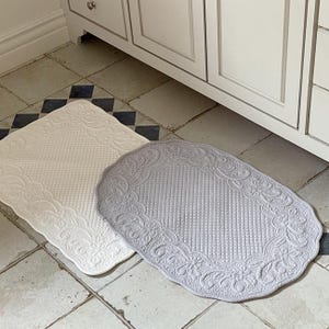 Puede incluir: Dos alfombras de baño decorativas. Una es rectangular y de color crema, la otra es ovalada y gris. Ambas tienen un diseño de borde floral y una textura acolchada. Se colocan sobre un suelo embaldosado cerca de un mueble blanco.