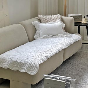 May include: A white, quilted sofa cover with a scalloped edge and floral pattern, placed on a beige sofa. A white pillow with ruffled edges and a smaller, textured pillow are also on the sofa. The room has a neutral colour palette.