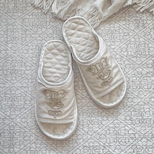 以下が含まれることがあります： キルト加工されたインソールと装飾的な刺繍が施された、オフホワイトのオープントゥスリッパのペア。スリッパは柔らかく、豪華な外観で、屋内での使用を目的としています。刺繍は、華やかな花柄のデザインが特徴です。