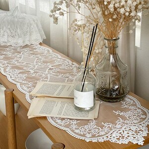 Op de afbeelding: Een witte kanten tafelloper met een delicaat bloemmotief ligt op een houten plank. Een heldere glazen fles met riet en een vintage vaas met gedroogde bloemen staan ook op de plank. Twee open boeken liggen op de plank.