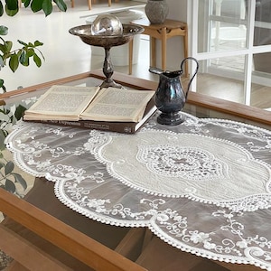 May include: A decorative table setting featuring a vintage silver-toned serving dish with a glass sphere, a small dark pitcher, and an open book. A white lace runner with floral and scalloped edges adds a delicate touch to the glass table.