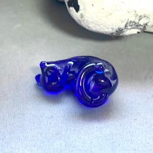 May include: A small, cobalt blue glass cat figurine. The cat is curled up, with its tail wrapped around its body. The glass is smooth and reflective, with a glossy finish. The cat is lying on a light grey surface.