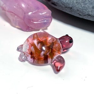 May include: A miniature glass turtle ornament. The turtle has a translucent pink head and flippers, and a shell of mixed pink, orange, and white glass. The turtle's eyes are small black dots. A pink glass sculpture is in the background.