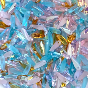 May include: A close-up shot of a collection of teardrop-shaped beads in various colors. The beads are primarily shades of blue, pink, and gold, with some iridescent and translucent elements. The beads are scattered, creating a textured and colorful display.