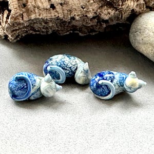 Pode incluir: Três pequenas figuras de ratos de vidro artesanais. Cada rato é principalmente azul com marmoreio branco, e tem uma cauda enrolada e orelhas pequenas. Os ratos estão dispostos sobre uma superfície cinzenta, com um pedaço de madeira e uma pedra no fundo.