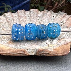 May include: Four blue glass beads with silver-colored dots are displayed on a textured, light brown surface. The beads have a rounded, cylindrical shape and vary slightly in shade of blue.