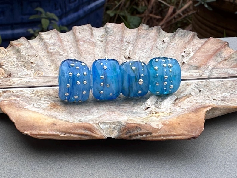 May include: Four blue glass beads with silver-colored dots are displayed on a textured, light brown surface. The beads have a rounded, cylindrical shape and vary slightly in shade of blue.