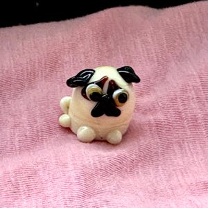 Puede incluir: Una pequeña figura de un pug de color crema con detalles negros en las orejas, la nariz y los ojos. El pug está sentado sobre una superficie rosa texturizada. La figura tiene un cuerpo redondeado y pies pequeños y redondos.