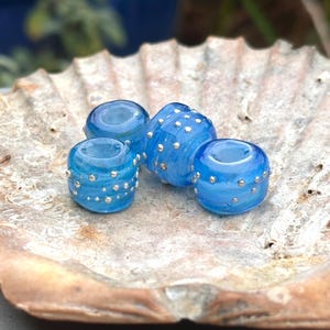 May include: Four translucent blue glass beads with silver accents. The beads are cylindrical and have a dotted pattern. They are displayed on a beige seashell, creating a contrast of textures and colors. The beads are likely for jewelry making.