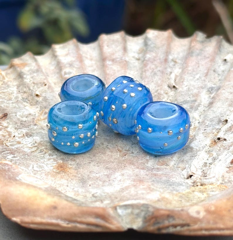 May include: Four translucent blue glass beads with silver accents. The beads are cylindrical and have a dotted pattern. They are displayed on a beige seashell, creating a contrast of textures and colors. The beads are likely for jewelry making.