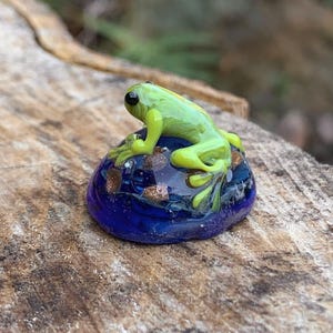May include: A small glass sculpture of a green frog sitting on a blue and gold glass rock.