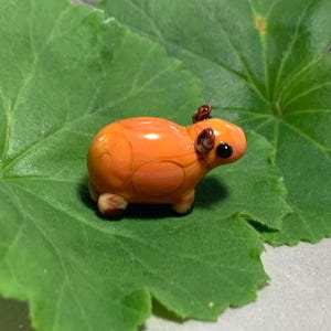 Może przedstawiać: Mała, pomarańczowa szklana figurka kapibary z ciemnymi oczami i małymi uszami. Figurka spoczywa na dużym, zielonym liściu. Kapibara ma błyszczące wykończenie i brązowe akcenty.