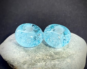 May include: Two light blue glass beads with a hole in the center. The beads have a bubbly texture.