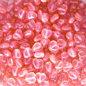 May include: A close-up of a collection of translucent pink heart-shaped beads. Each bead has a glossy finish and a white stripe. The beads are scattered, creating a textured surface.