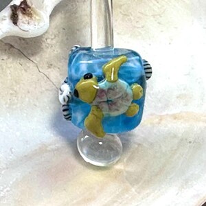 May include: A handmade glass bead with a sea turtle design. The bead has a translucent blue background with a yellow and pink turtle. The bead is mounted on a clear glass rod and base. The bead is approximately 2 cm in height.