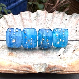 May include: Four blue glass beads with silver accents are displayed on a seashell. The beads have a rectangular shape and a translucent quality, with the silver accents appearing as small, irregular dots. The beads are a vibrant blue color.