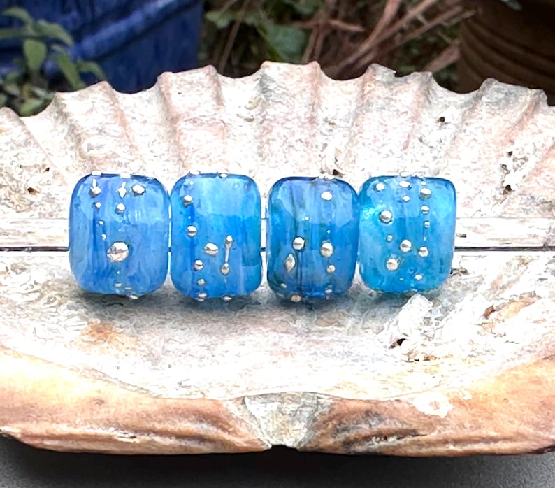 May include: Four blue glass beads with silver accents are displayed on a seashell. The beads have a rectangular shape and a translucent quality, with the silver accents appearing as small, irregular dots. The beads are a vibrant blue color.