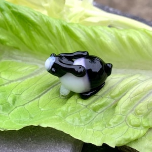 Może przedstawiać: Mała, ręcznie wykonana szklana figurka pandy w czarno-białej kolorystyce. Panda leży na dużym, zielonym liściu sałaty. Ciało pandy jest białe, z czarnymi akcentami na głowie, ramionach i stopach.