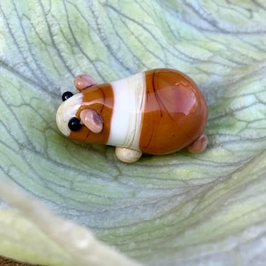 May include: A glass guinea pig ornament. The small animal has a brown and white body with black eyes and pink ears. The figurine is displayed on a green leaf.