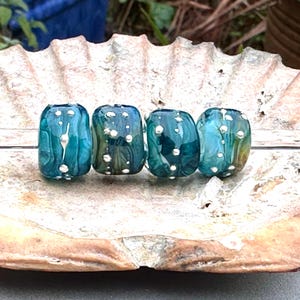 Puede incluir: Cuatro cuentas de vidrio color turquesa con puntos blancos, exhibidas sobre una concha. Las cuentas tienen un efecto veteado con tonos de azul, verde y amarillo. Cada cuenta mide aproximadamente 2 cm de largo.
