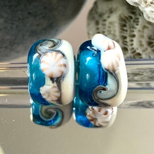 Op de afbeelding: Twee blauwe en witte glazen kralen met een schelpdesign. De kralen hebben een swirl-patroon en zijn versierd met witte schelpen.