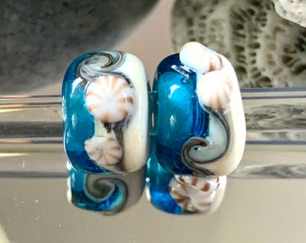 May include: Two blue and white glass beads with a seashell design. The beads have a swirl pattern and are decorated with white seashells.