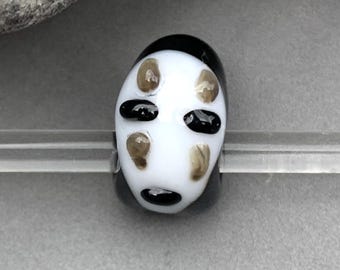May include: A white glass bead with a black and brown No Face character from the movie Spirited Away.