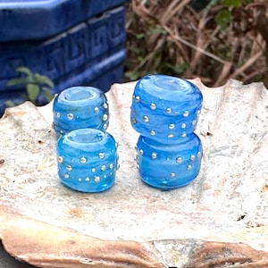 May include: Four translucent blue glass beads with silver accents. Two beads are stacked, and two are separate. The beads are cylindrical with a hole through the center. The beads are on a light-colored, textured surface.