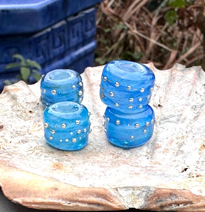 May include: Four translucent blue glass beads with silver accents. Two beads are stacked, and two are separate. The beads are cylindrical with a hole through the center. The beads are on a light-colored, textured surface.