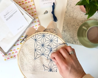 May include: A close-up of a person stitching a blue thread onto a white fabric with a geometric pattern. The fabric is stretched in a wooden embroidery hoop. The text "The Wildest One" is visible on a card in the background.