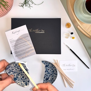 K&ouml;nnte beinhalten: Ein Kintsugi-Reparaturset von The Wildest One mit einer schwarzen Box, Anleitungskarte, Goldpulver, Klebstoff, einem feinen Pinsel, h&ouml;lzernen Mischst&auml;bchen und einer blau-wei&szlig;en, floralen Keramikschale, die in zwei Teile zerbrochen ist.
