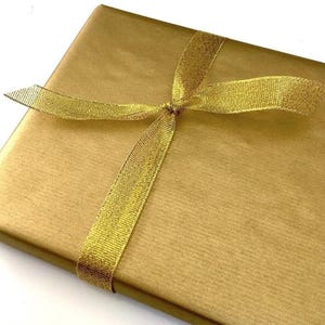 K&ouml;nnte beinhalten: Eine rechteckige Geschenkbox, eingewickelt in glattes, metallisch goldenes Papier. Das Paket ist mit einem schimmernden, durchscheinenden goldenen Band umwickelt, das ein einfaches Kreuzmuster bildet und mittig verknotet ist.