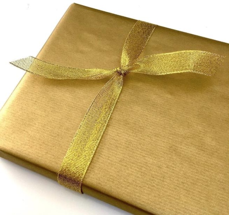 K&ouml;nnte beinhalten: Eine rechteckige Geschenkbox, eingewickelt in glattes, metallisch goldenes Papier. Das Paket ist mit einem schimmernden, durchscheinenden goldenen Band umwickelt, das ein einfaches Kreuzmuster bildet und mittig verknotet ist.