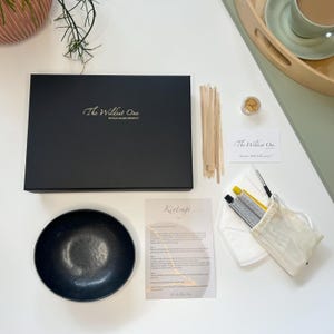 May include: A Kintsugi kit containing a black bowl, gold paint, wooden sticks, a small jar of gold flakes, a white face mask, a white cloth bag, and instructions. The kit is packaged in a black box with the text "The Wildest One" on the top.