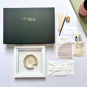 May include: A Kintsugi repair kit featuring a large dark green box labelled The Wildest One, a white framed ceramic piece with a gold-filled crack, wooden mixing sticks, a black brush, gold powder, two paint tubes, a mask, and instruction cards.