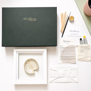 May include: A Kintsugi repair kit featuring a large dark green box with gold text, a white frame containing a small ceramic dish with a gold-filled crack, wooden mixing sticks, a brush, gold powder, two paint tubes, a mask, and instruction cards.